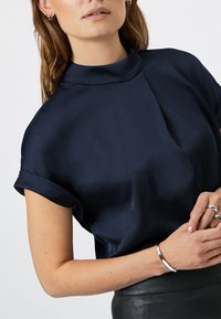 Navyblauwe zijden blouse met een hoge kraag, korte opgerolde mouwen en een gladde textuur. Geaccentueerd door een zilveren armband om een pols.