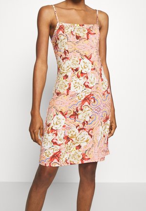 Frau in einem ärmellosen, knielangen Kleid mit dünnen Trägern und einem rosa, weißen und roten floralem sowie paisley-muster.