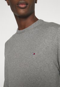 Homme portant un pull en maille gris avec un petit logo rectangulaire rouge, blanc et bleu sur la poitrine, sur un fond uni.