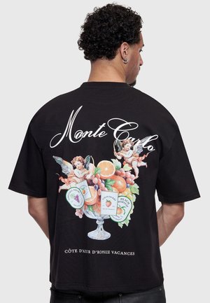 Bărbat purtând un tricou negru cu textul „Monte Carlo”, îngerași și o grafică cu un bol de fructe pe spate.