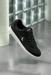 Black leather sneaker with white rubber sole, featuring a round toe, laces, and a logo on the side. Smooth texture and minimal design.