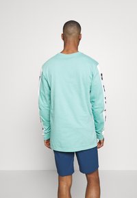 Světle modré tričko s dlouhým rukávem a bílými rukávy, na kterých je černý text. Tkanina vypadá měkce a střih je volný. Celé je doplněno námořnickými šortkami.