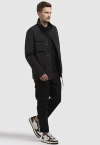 Svart jacka med flera framfickor, knappknäppning fram och hög krage, i kombination med svarta cargobyxor och svart-vita sneakers.