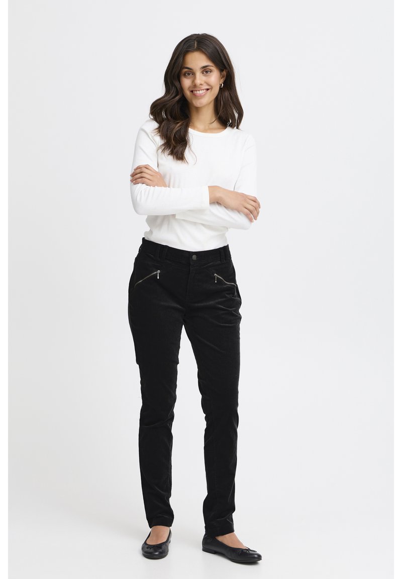 Fekete kordbársony nadrág két cipzáras elülső zsebbel, fehér hosszú ujjú inggel párosítva. A modell összefont karokkal áll, mosolyogva.
