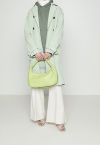 Persona con abrigo verde menta, sudadera verde oliva, pantalones anchos blancos, sandalias con tiras, sosteniendo un bolso verde lima frente a un fondo plano.