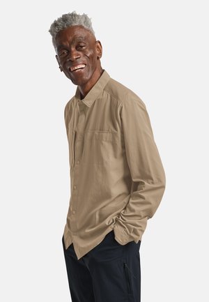 Uomo anziano con capelli grigi che indossa una camicia marrone chiaro abbottonata e pantaloni neri, sorride con una mano in tasca davanti a uno sfondo semplice.