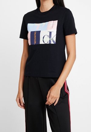 Femme portant un t-shirt noir Calvin Klein avec un logo coloré en bloc et un pantalon noir avec des bandes rouges sur les côtés, debout sur fond blanc.