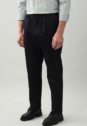 Homme portant un pantalon noir sur mesure, des chaussures noires à lacets et une chemise claire à manches longues avec des poignets retroussés.