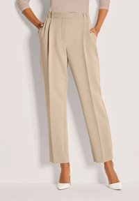 Vrouw in beige high-waisted geplooide broeken en witte puntige hoge hakken, staand met haar handen in haar zakken tegen een lichte achtergrond.