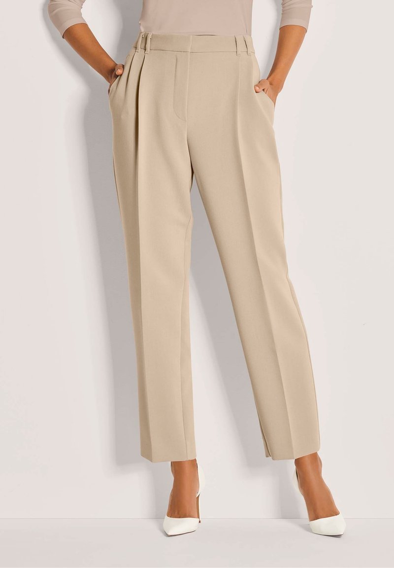 Vrouw in beige high-waisted geplooide broeken en witte puntige hoge hakken, staand met haar handen in haar zakken tegen een lichte achtergrond.