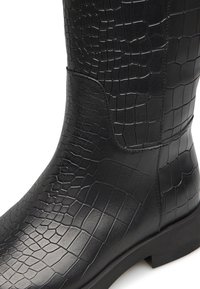 Stivale alto in pelle nera con texture coccodrillo, dettagli con cuciture curve e una robusta suola piatta.