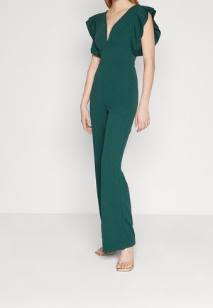 Kvinde iført en mørkegrøn jumpsuit med flæseærmer og dyb V-udskæring, kombineret med gyldne sandaler med åben tå, stående mod en ensfarvet hvid baggrund.