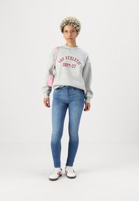Grå hettegenser med "GAP ATHLETIC DEPT-27" i rødt skrifttype. Slanke blå jeans, hvite joggesko med røde striper, og en liten rosa håndveske.