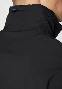 Veste soft-shell noire avec un col haut, dotée d'un cordon de serrage bleu et de détails de couture subtils, présentée en vue rapprochée.