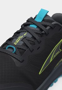 Κοντινό πλάνο ενός μαύρου αθλητικού παπουτσιού με κορδόνια με σχέδιο, μπλε θηλιά για να το τραβάς, πράσινο λογότυπο και το κείμενο «Altra Lone Peak» στη γλώσσα του παπουτσιού.