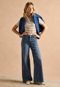 Džínsy s širokými nohavicami v kombinácii s bielo-hnedou prúžkovanou polokošeľou a modrou denimovou vestou prehodenou cez ramená. Biele topánky.