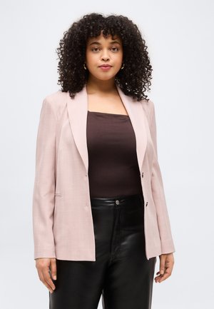 Woman with curly hair wearing gold hoop earrings, a light pink blazer, dark brown top, and black leather pants, standing against a white background.