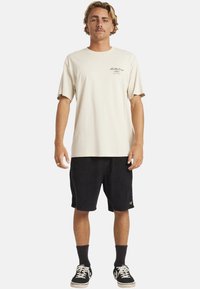 Crème T-shirt met een klein logo op de borst, gecombineerd met zwarte corduroy shorts en zwarte sneakers met witte zolen en veters.