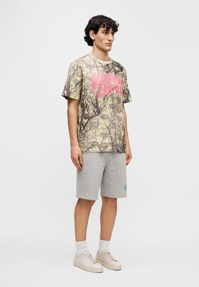 Βαμβακερό T-shirt με καμουφλάζ μοτίβο σε αποχρώσεις του πράσινου και καφέ· διαθέτει έντονο ροζ κείμενο. Συνδυάζεται με γκρι σορτς και ανοιχτόχρωμα αθλητικά παπούτσια.