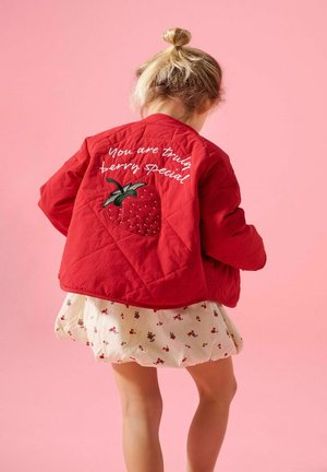 Barn med blondt hår iført en rød jakke med en påsydd jordbær og teksten «You are truly berry special», over et kremfarget skjørt med små røde blomster.