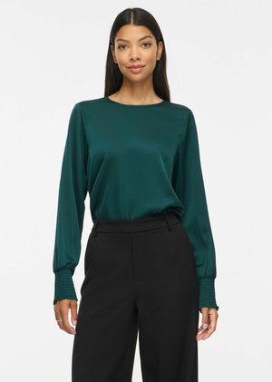 Femme portant un chemisier en satin vert foncé à manches longues avec poignets froncés et un pantalon noir taille haute, debout devant un fond blanc.