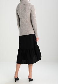 Béžový pletený svetr s vysokým límcem, zkombinovaný s černými, vrstevnatými šaty midi délky. Hladká textura, přiléhavý střih nahoře, rozšířený dole.