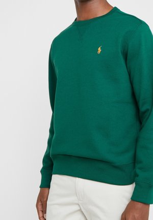 Mann trägt einen grünen Rundhalspullover mit kleinem gelbem besticktem Pferdelogo auf der Brust und offwhite Hose.
