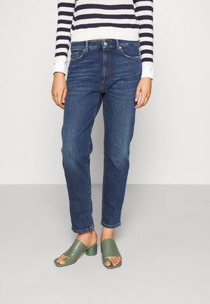 Persoon draagt donkerblauwe spijkerbroek, groene open teen sandalen met blokhak, en een wit shirt met marineblauwe strepen, staand op een lichtgrijze vloer.