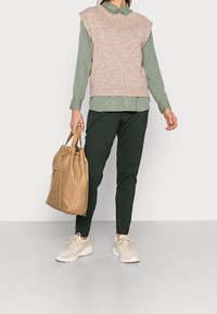 Weste in bunten Farben, kombiniert mit einem hellgrünen Hemd, dunkelgrünen Hosen und einer beigen Tragetasche mit Kordelzug. Die Sneakers sind hell und haben ein Mesh-Design.