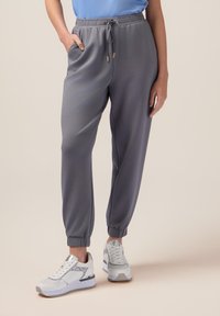 Graue Jogginghose mit elastischem Bund und Kordelzug, ausgestattet mit Seitentaschen und gerippten Bündchen. Kombiniert mit weißen Sportschuhen.