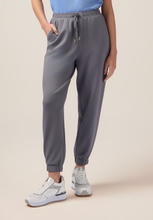 Graue Jogginghose mit elastischem Bund und Kordelzug, ausgestattet mit Seitentaschen und gerippten Bündchen. Kombiniert mit weißen Sportschuhen.