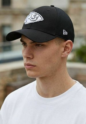 Junger Mann, der eine schwarze Baseballkappe mit weißem Logo und ein weißes T-Shirt mit Rundhalsausschnitt trägt, nach links blickend vor einem unscharfen Hintergrund im Freien.