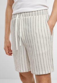 Pantalones cortos de color claro, tejidos, con rayas verticales en gris, cinturilla elástica, cordón ajustable y bolsillos laterales. Textura suave.