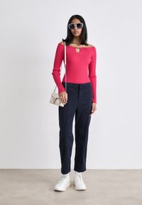 Top rosa a costine con spalle scoperte e dettaglio a chiave, abbinato a pantaloni blu navy e scarpe da ginnastica bianche. Gli accessori includono una piccola borsa a tracolla di colore chiaro.