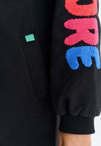 Czarna rękaw z kolorowymi, teksturowanymi literami „ARE” w kolorach czerwonym, różowym i niebieskim na tkaninie, obok czarnej kieszeni z małą zieloną metką.