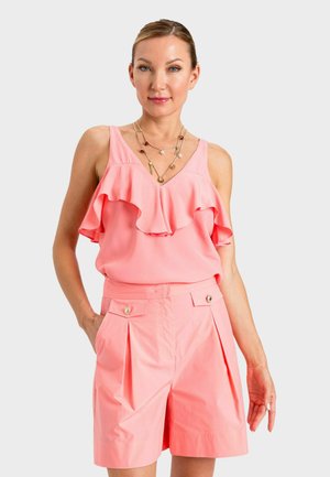 Donna che indossa un top senza maniche rosa corallo con volant e pantaloncini a vita alta con tasche con bottoni, in piedi con una mano in tasca.