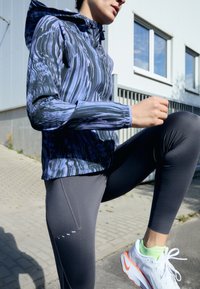 Osoba ubrana w wzorzystą kurtkę i ciemne legginsy unosi jedno kolano na zewnątrz, a na utwardzonej ścieżce widoczne są białe i pomarańczowe buty do biegania.