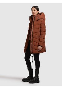 Brun pufferjakke med hætte, vandret quiltet design og lynlås foran. SAmmen med sorte leggings og kraftige sorte støvler.