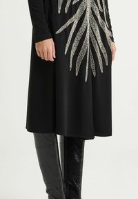 Schwarzes Kleid mit langen Ärmeln, verziert mit metallic-silbernem Blattmuster. Kombiniert mit kniehohen schwarzen, strukturierten Stiefeln, die eine schlanke Silhouette betonen.