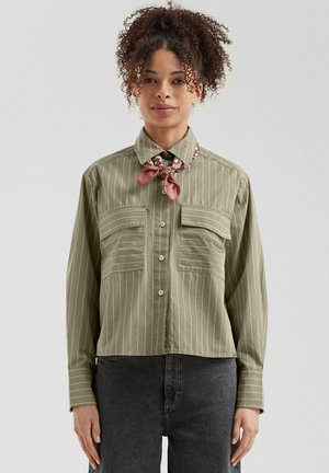 Vrouw in een olijfgroene gestreepte blouse met knopen, twee borstzakken en een rode met patroon geknoopte sjaal, staande tegen een effen achtergrond.