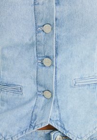 Ljusblå denimväst med silverfärgade metallknappar, två bröstfickor och en svagt V-formad nederkant. Texturen är vävd med synlig sömnad.