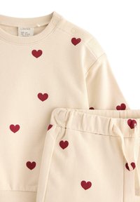 Ljust beige sweatshirt och matchande byxor, båda med röda hjärtan. Sweatshirten har tryckknappar vid axeln. Mjuk material.