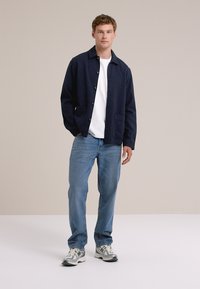 Man staat zelfverzekerd in een marineblauwe jas boven een wit shirt, gecombineerd met blauwe jeans en sneakers, tegen een neutrale achtergrond.