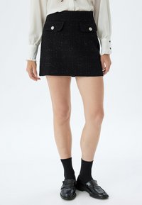 Mini-jupe en tweed noir avec deux poches avant, accents de boutons argentés et un motif texturé. Associée à des mocassins noirs et des chaussettes.
