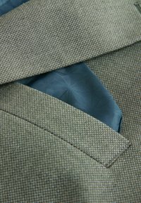 Nahaufnahme einer strukturierten beigefarbenen Blazer-Tasche, die ein gefaltetes, blau gemustertes Seidentaschentuch hält.