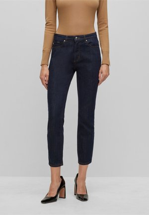 Personne portant un jean court bleu foncé, un haut beige à manches longues et des chaussures à talons noires se tenant devant un fond uni.