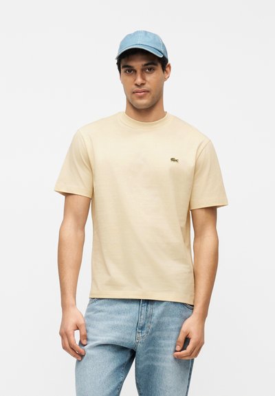 Jeune homme portant un t-shirt beige à manches courtes avec un petit logo, un jean bleu clair et une casquette de baseball bleu clair, debout devant un fond blanc.