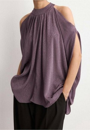 Kvinde iført løs lilla cold-shoulder bluse med plisseringer og sorte bukser, stående med den ene hånd i lommen mod en ensfarvet baggrund.