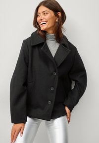 Cappotto in misto lana nero con ampio colletto a rever, quattro bottoni e tasche laterali, indossato sopra un dolcevita grigio e pantaloni metallici argento.
