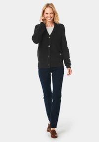 Schwarze gerippte Strickjacke mit V-Ausschnitt, Sechs-Knopf-Verschluss und langen Ärmeln, kombiniert mit dunkelblauen Hosen und braunen Slippern.